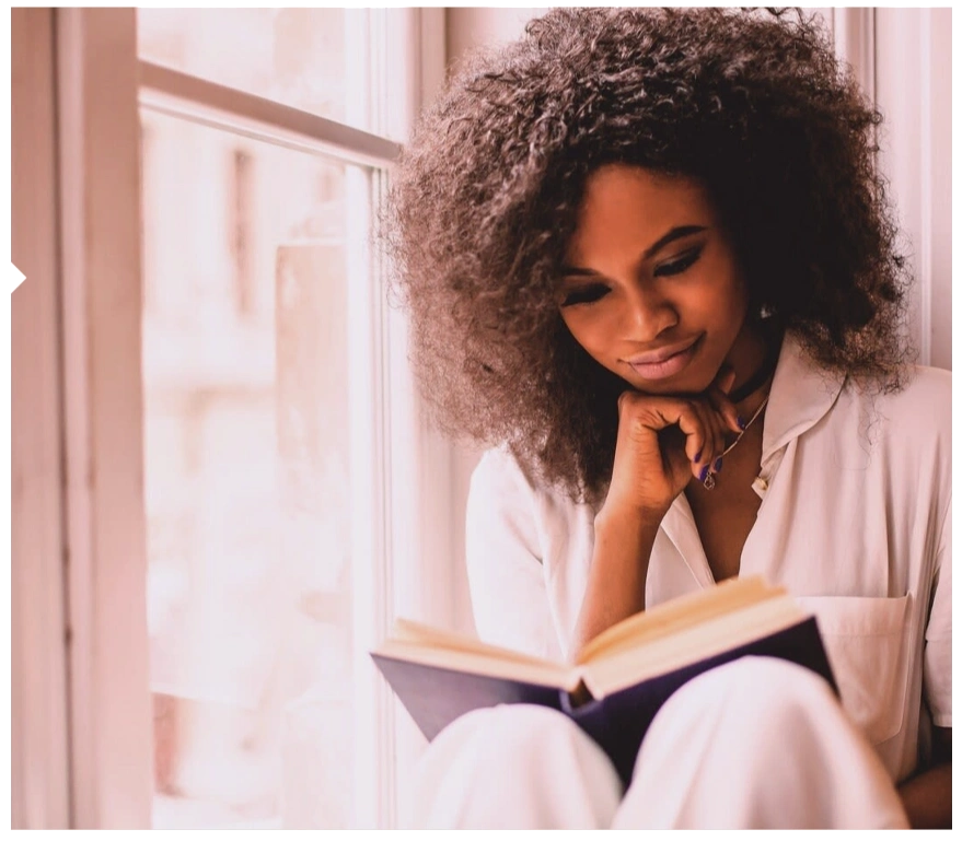 A woman with curly hair reading a book by a window.