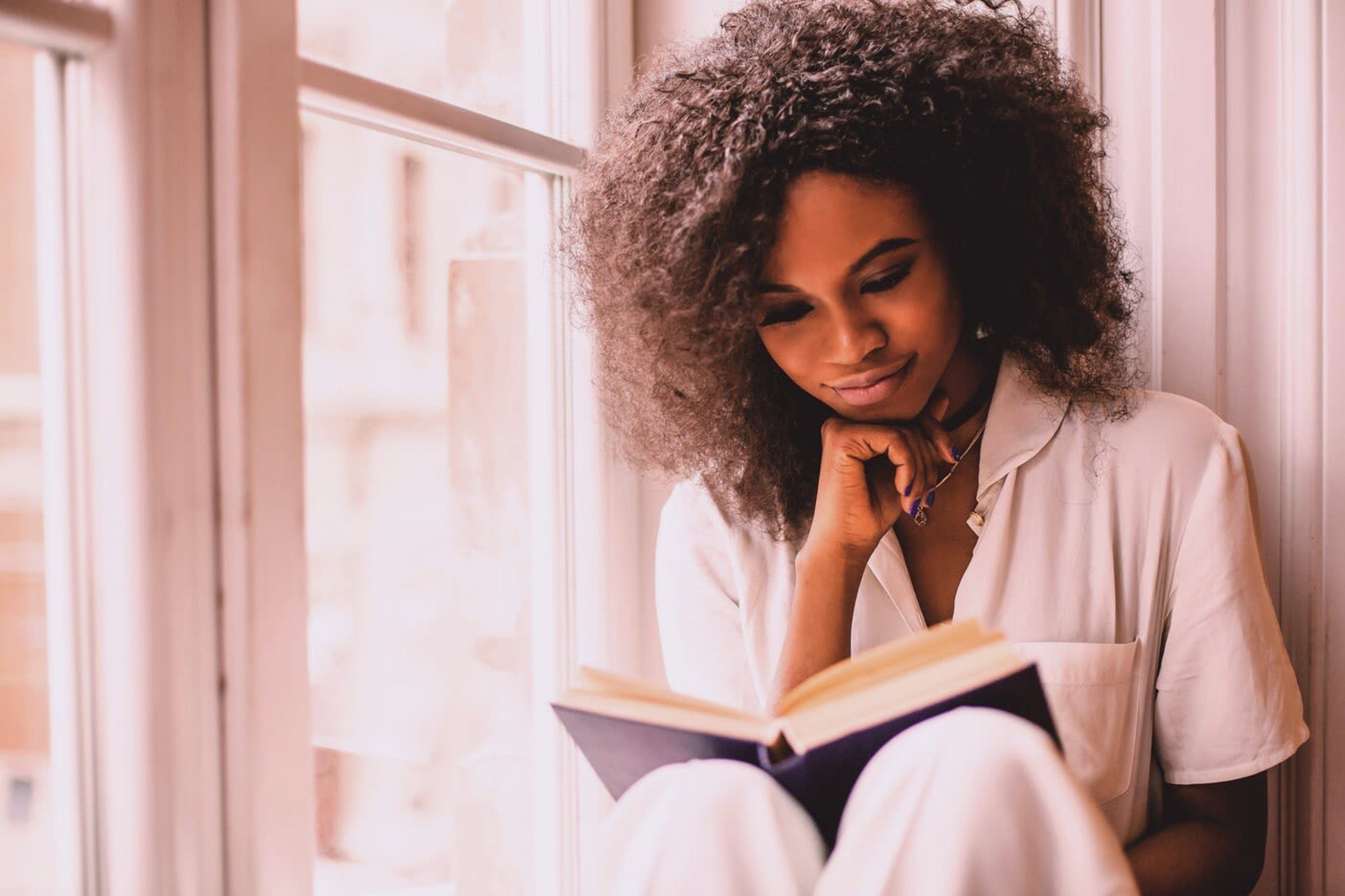 Woman reading a book by a sunlit window, looking thoughtful.